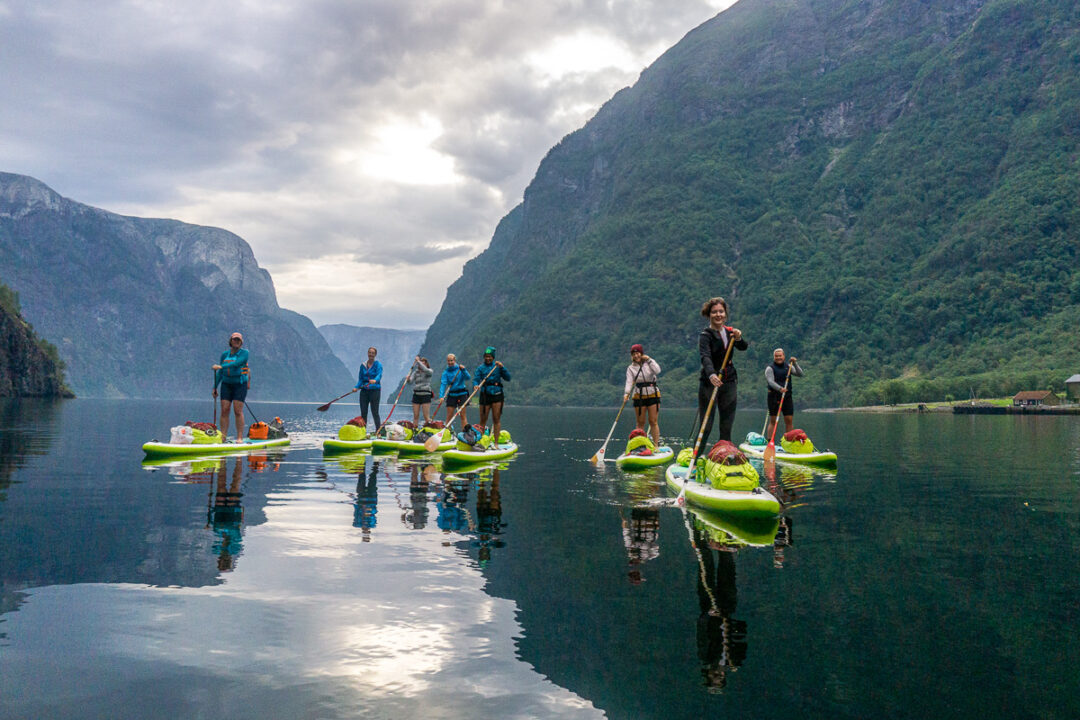 Women stand-up paddleboarding on a calm mountain lake with boats and steep green cliffs in the background