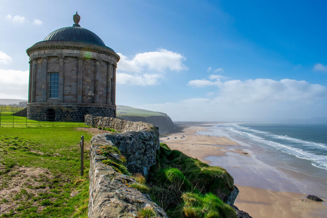 Coastal lighthouse on a grassy cliff overlooking a sandy beach and the sea, under a blue sky
