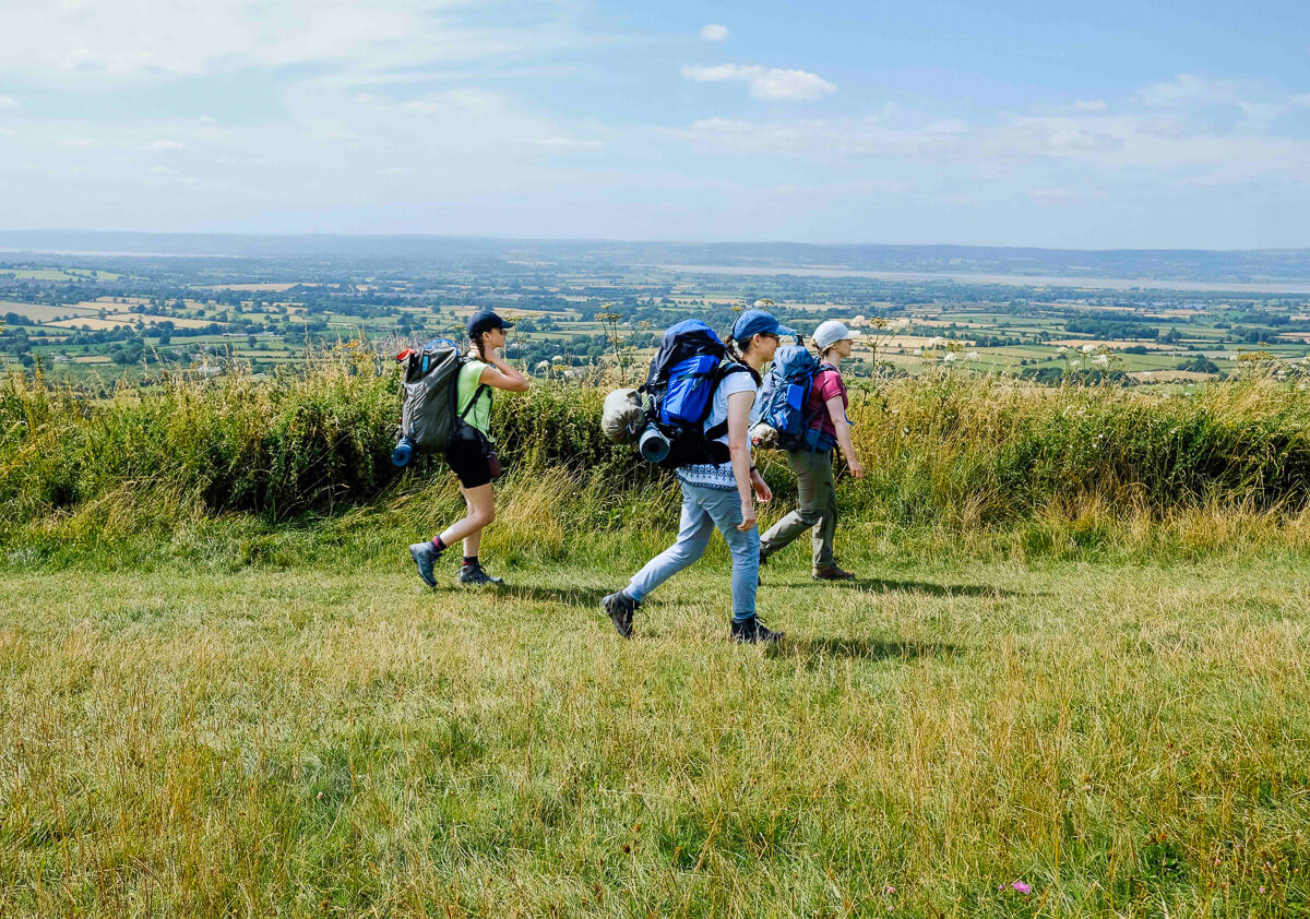 Women hiking with backpacks across a grassy hill overlooking countryside in Europe