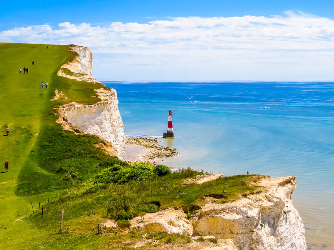 Clifftop hike past a lighthouse on dramatic white cliffs by the sea, with walkers along the coastal path