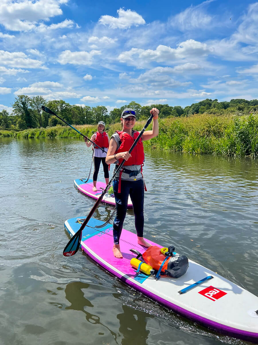 Women paddleboarding on a river, holding paddles on purple-and-white SUP boards under a blue sky