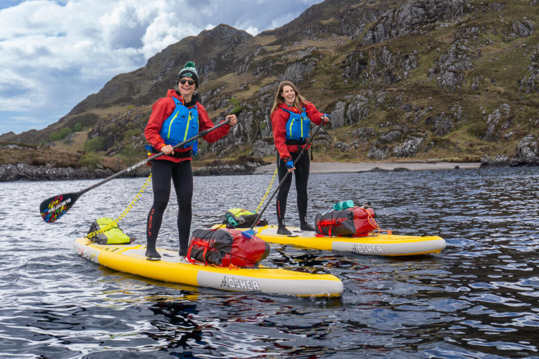 Women paddle boarding on yellow stand-up boards in a mountain lake landscape