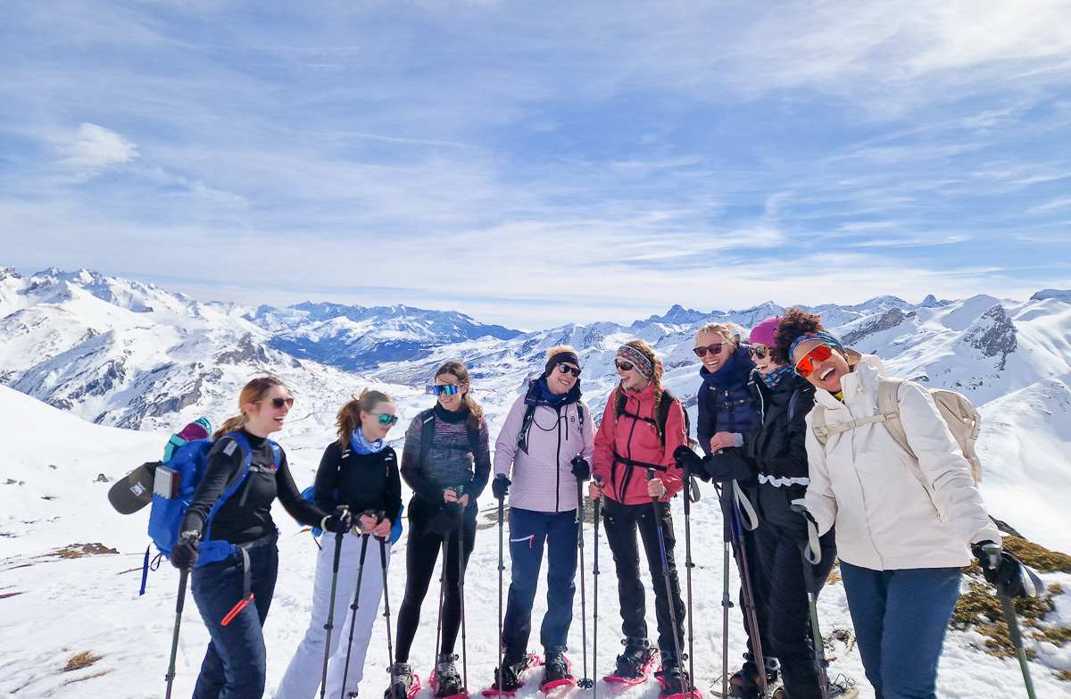Women trekking with poles on a snowy mountain ridge in Europe, surrounded by alpine peaks and blue sky.