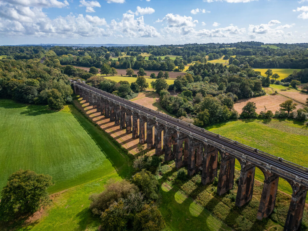 Historic stone viaduct bridge spanning green countryside fields and wooded hills under a blue sky