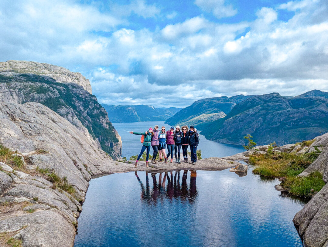 Group of women hiking on rocks beside a calm fjord in the mountains, with boats in the distance.