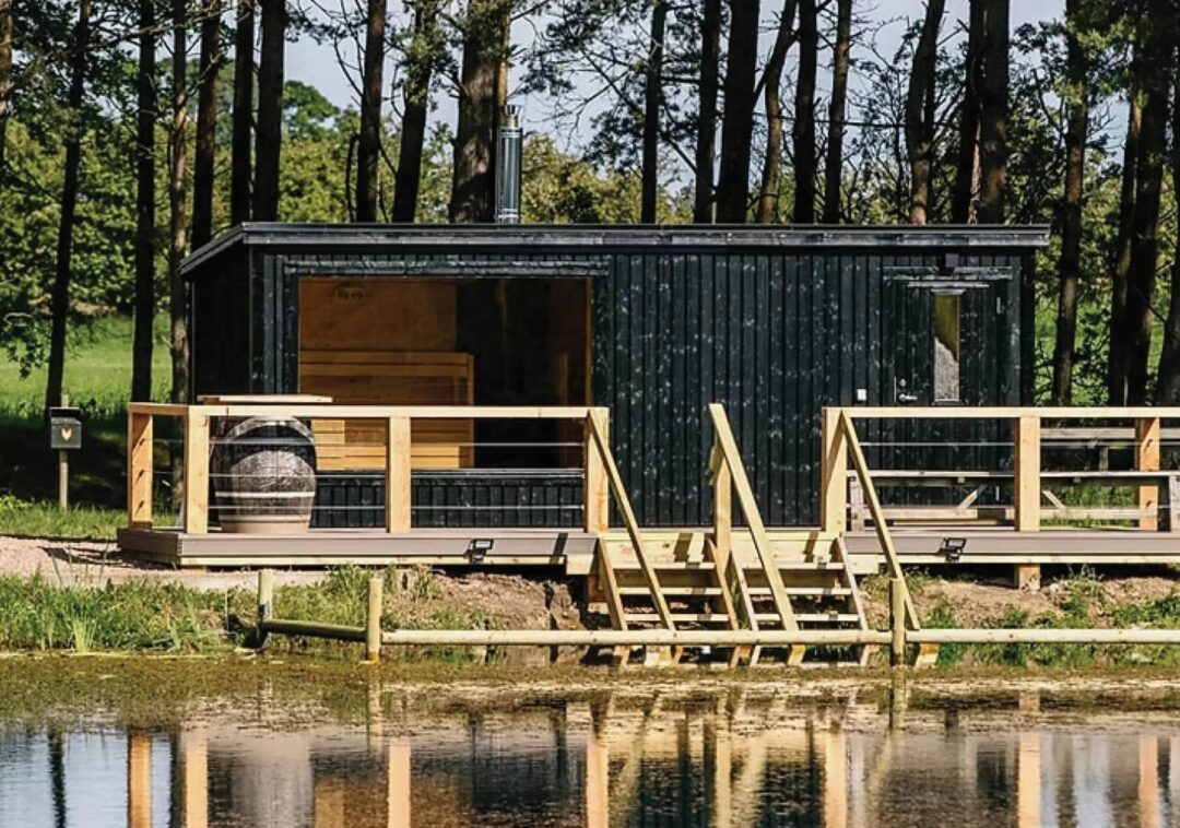 Wooden sauna cabin in a forest beside a pond, with outdoor steps and deck