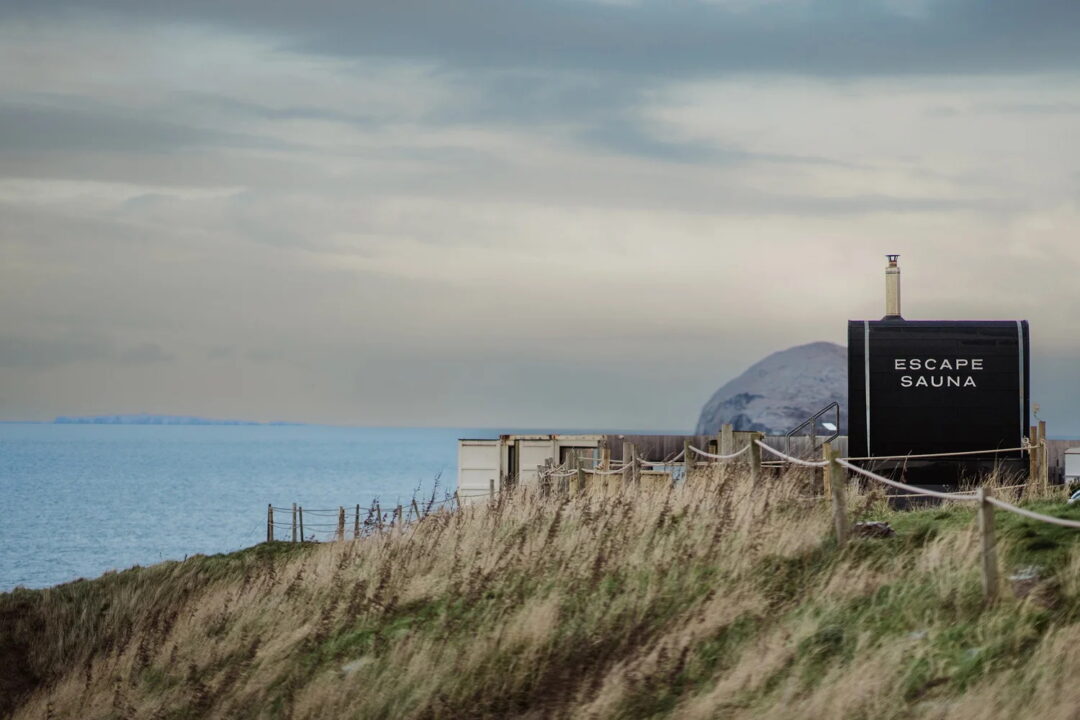 Escape Sauna by the sea in a sandy dune landscape, with a sign reading “ESCAPE SAUNA”