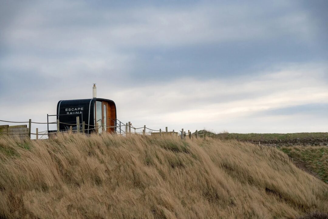 Sauna pod on seaside dunes with sign reading “ESCAPE RAIN”