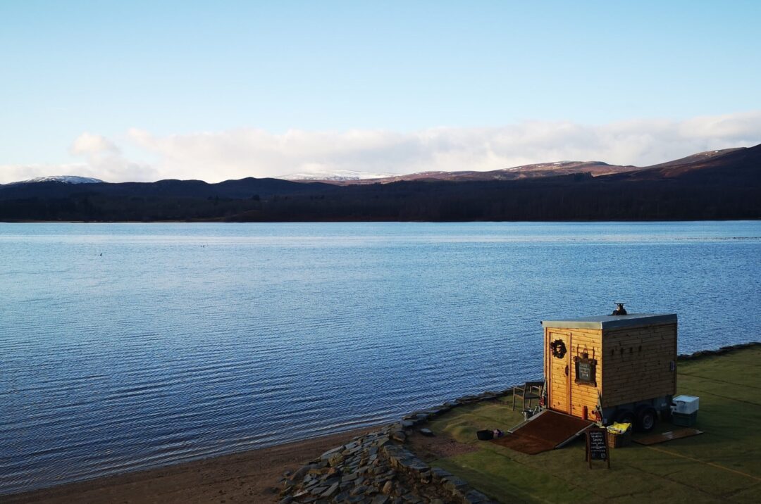 Wooden sauna cabin beside a calm loch under a blue sky