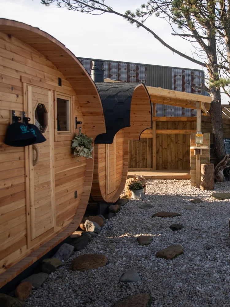 Wooden barrel sauna cabin outdoors with gravel courtyard and glass-panel building in the background