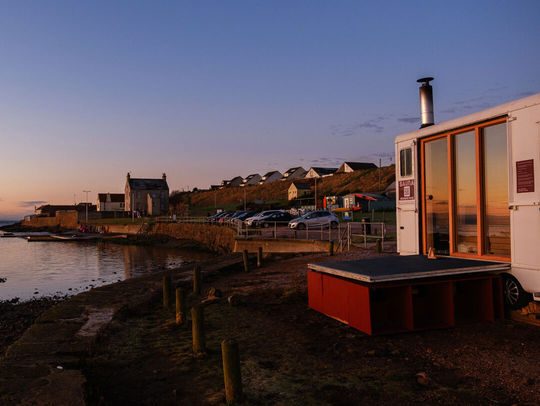 Outdoor wooden sauna cabin by a riverside at dusk, with houses and parked cars in the background
