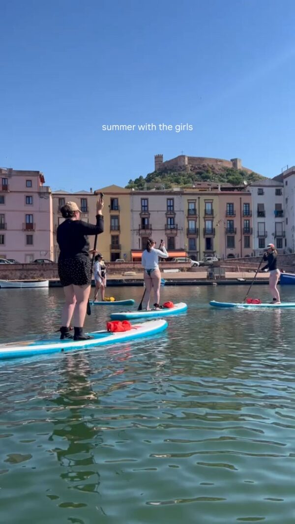 Women paddleboarding on blue boards by a harbour with a hilltop fortress; “summer with the girls”