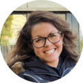 Smiling woman with glasses outdoors in a dark jacket.