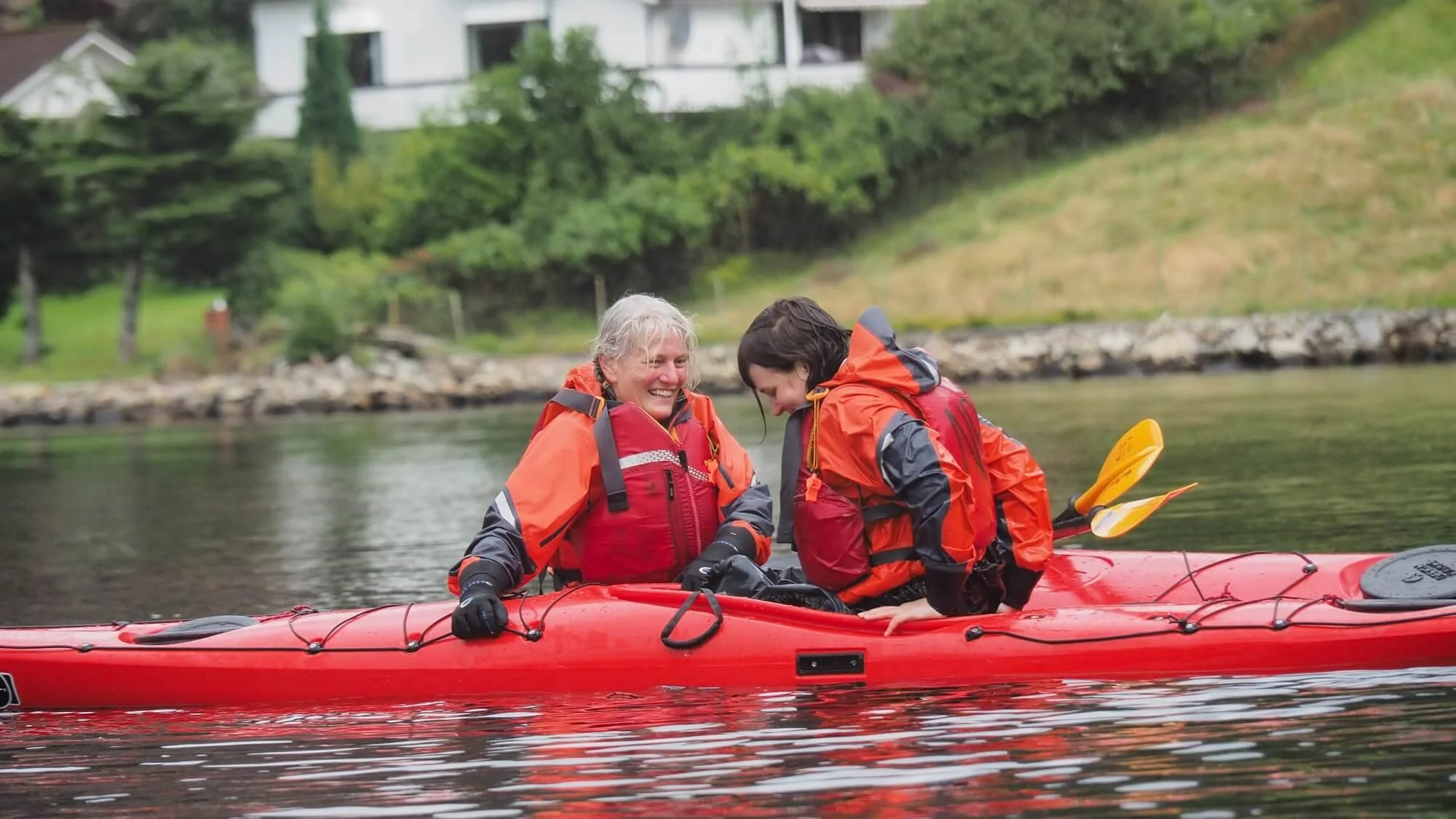 Women kayaking on a river, wearing life jackets in a red inflatable boat by a lakeside bank