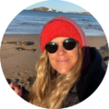 Woman wearing red beanie and sunglasses on a sandy beach by the sea with cliffs in the background