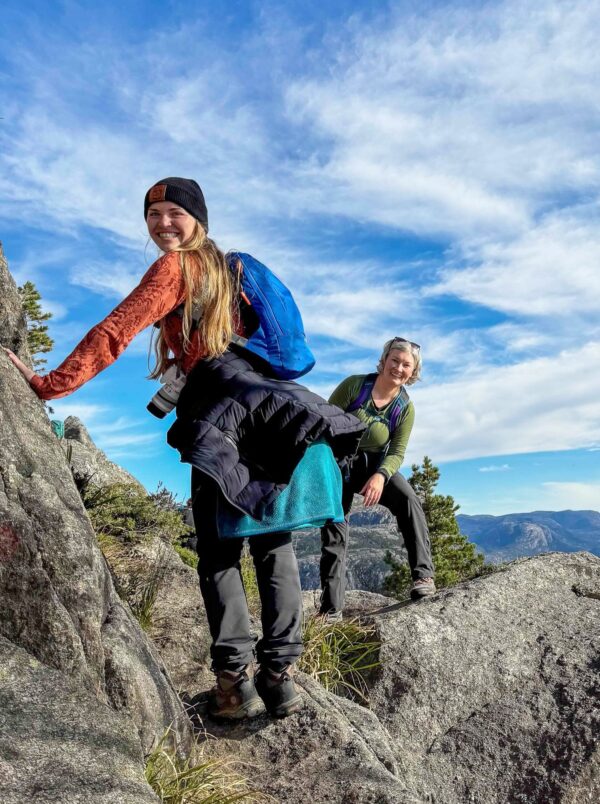 Two women on a hiking adventure, climbing rocks with backpacks under a blue sky and mountain views