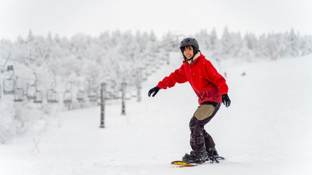 Woman snowboarding on a snowy slope, wearing a red jacket and helmet, with a ski resort in the background