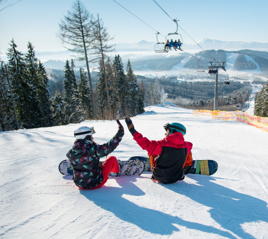 Women snowboarding on a snowy slope, with a chairlift overhead in a winter mountain landscape