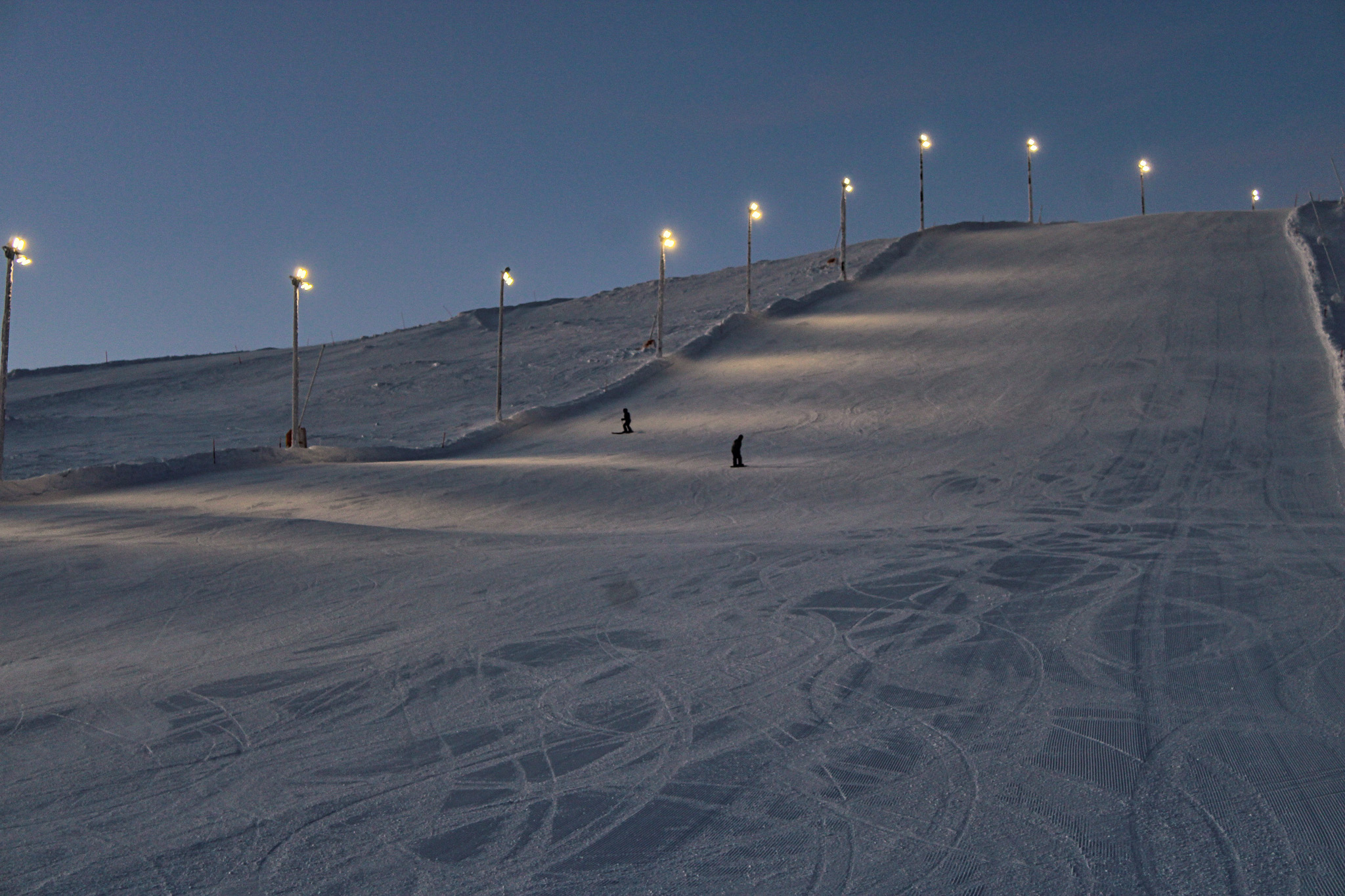 Skiers on a floodlit slope at dusk, with ski tracks and illuminated lamp posts.