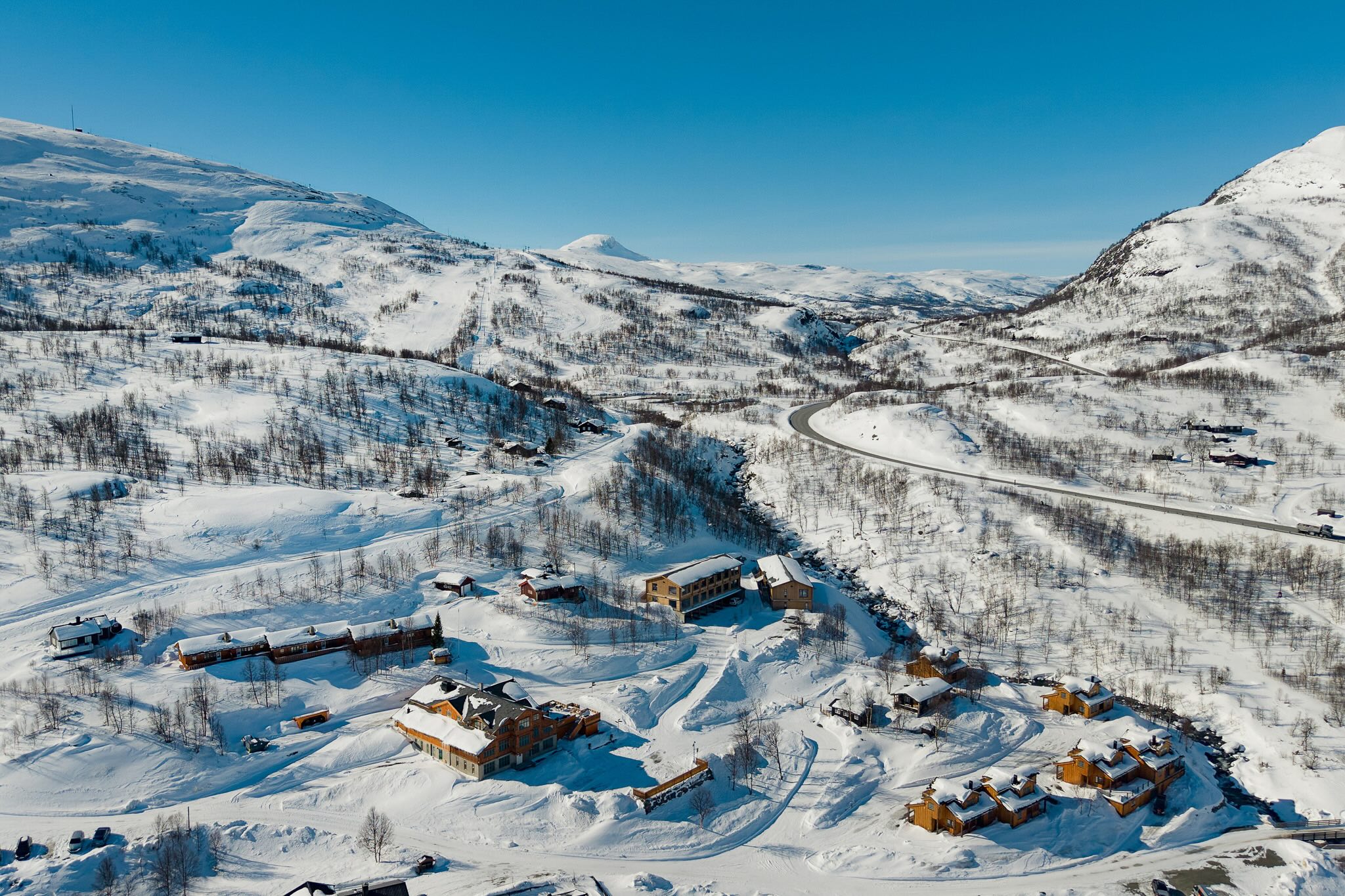 Aerial view of snowy Norwegian mountains with ski slopes, winding roads and cabins in winter.