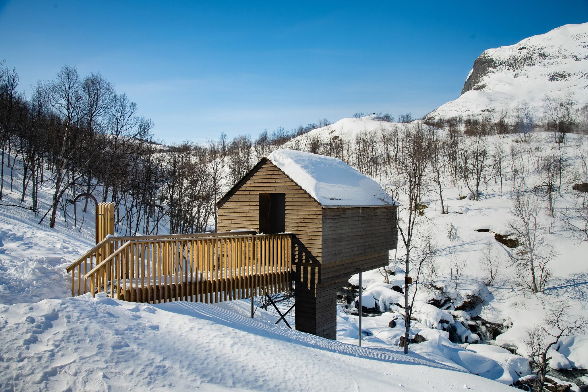Snow-covered wooden chalet in snowy Norwegian mountains, with chairlift-style support in foreground and blue sky above.