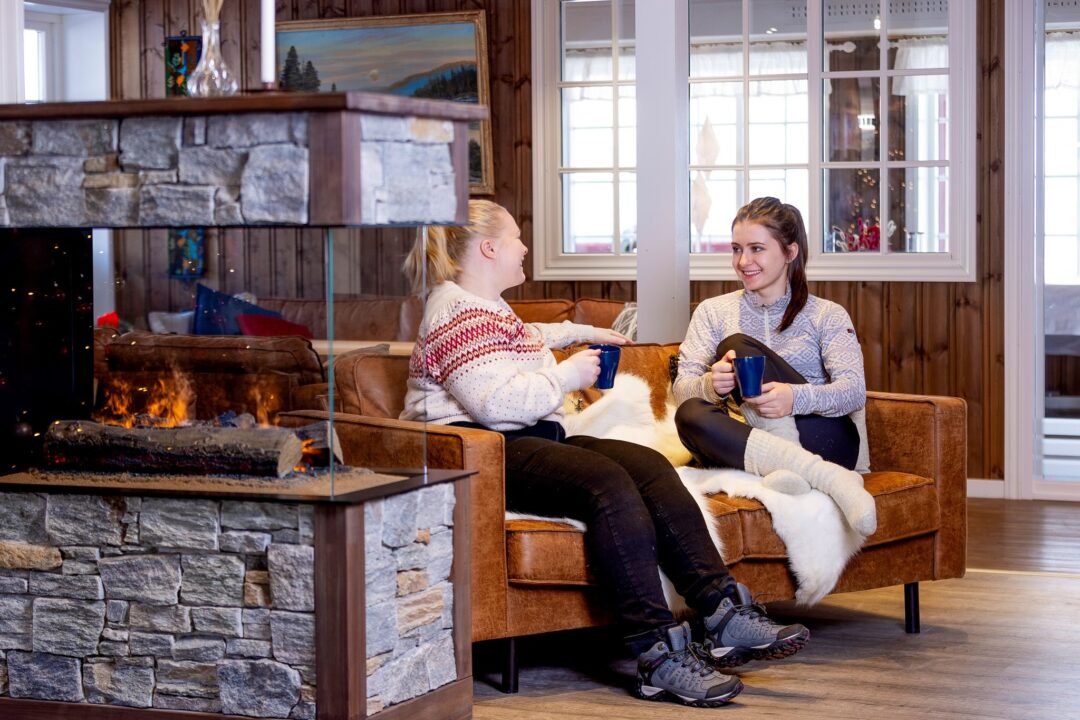 Two women relaxing by a roaring fireplace in a lodge, holding blue mugs and chatting indoors