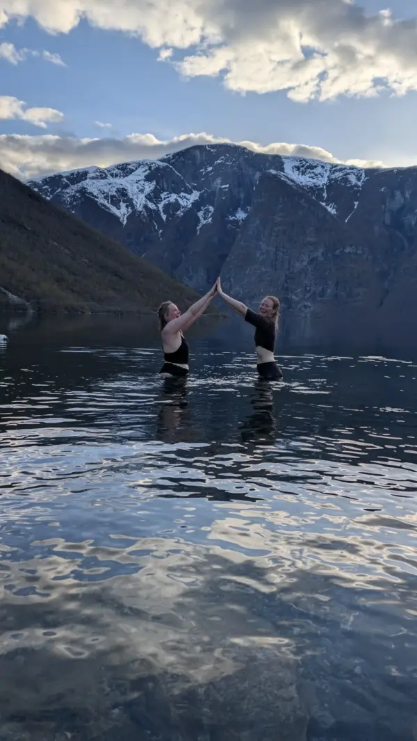 Two women wade in a fjord in Norway at sunset, with snow-capped mountains in the background