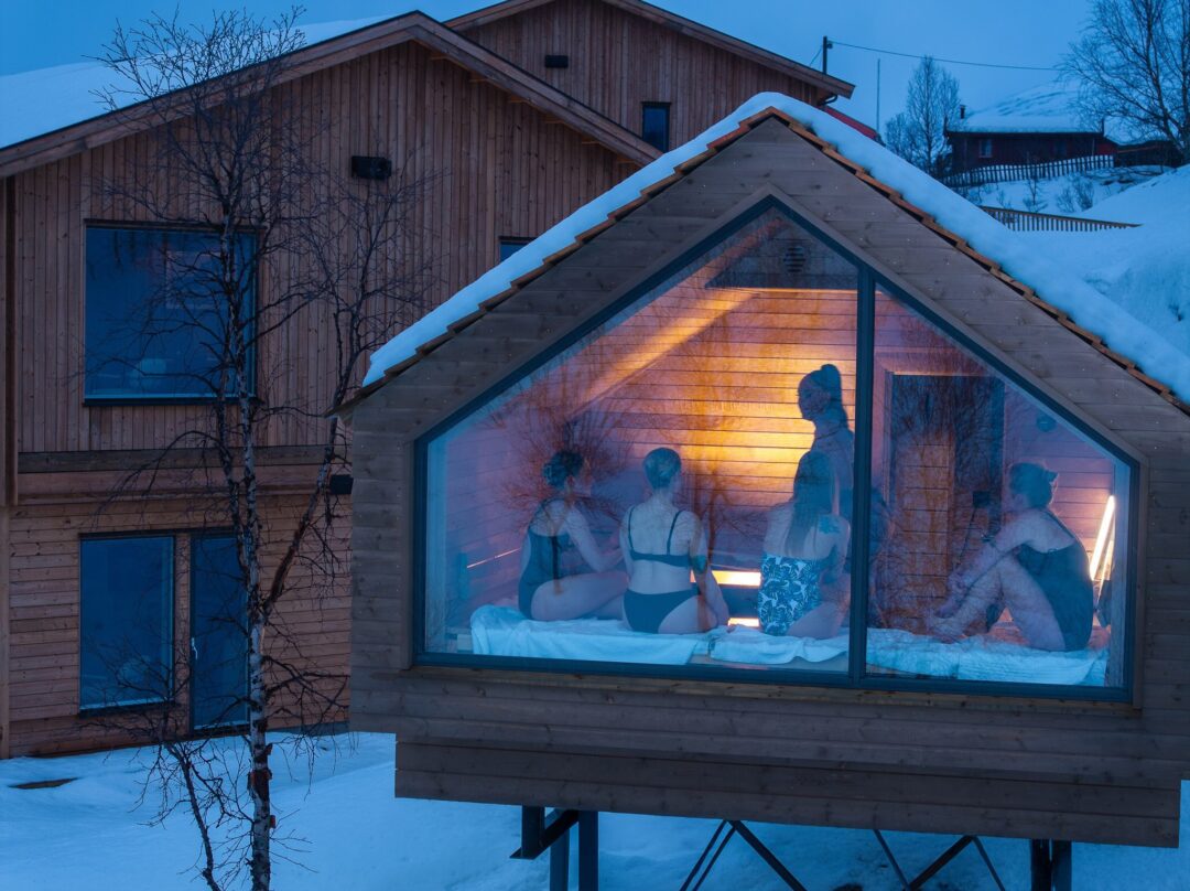 Women sitting on a window bench inside a snowy wooden chalet at dusk