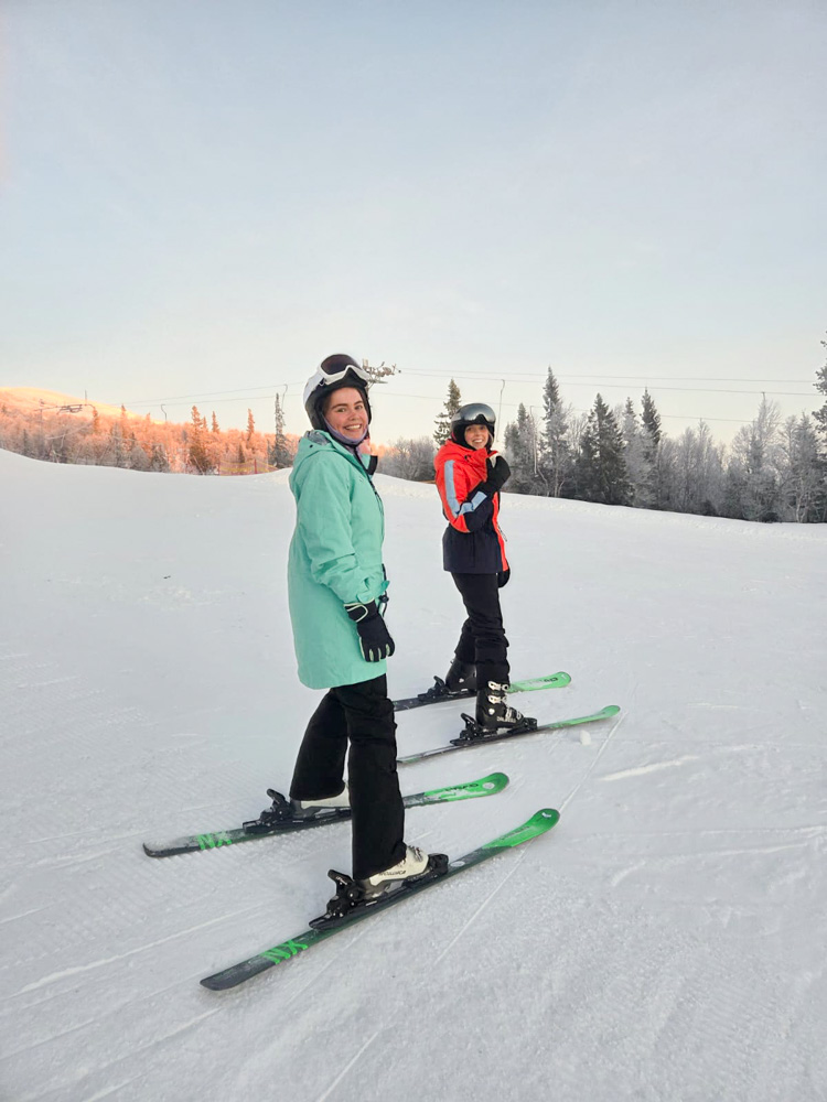 Women skate-ski on snow-covered slopes in Sweden at sunset, wearing skis and winter gear.