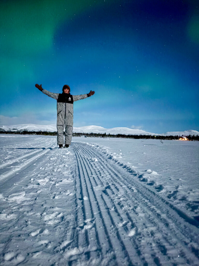 Woman cross-country skiing in snowy Norway under northern lights, standing with arms out on tracked snow