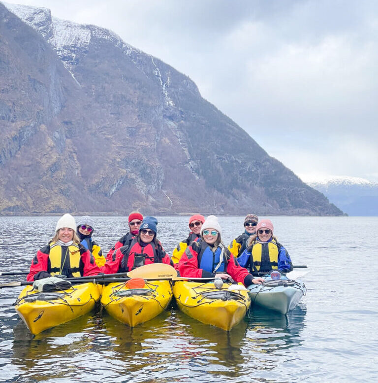 Women in yellow kayaks smiling on a Norway fjord with snow-dusted mountains behind